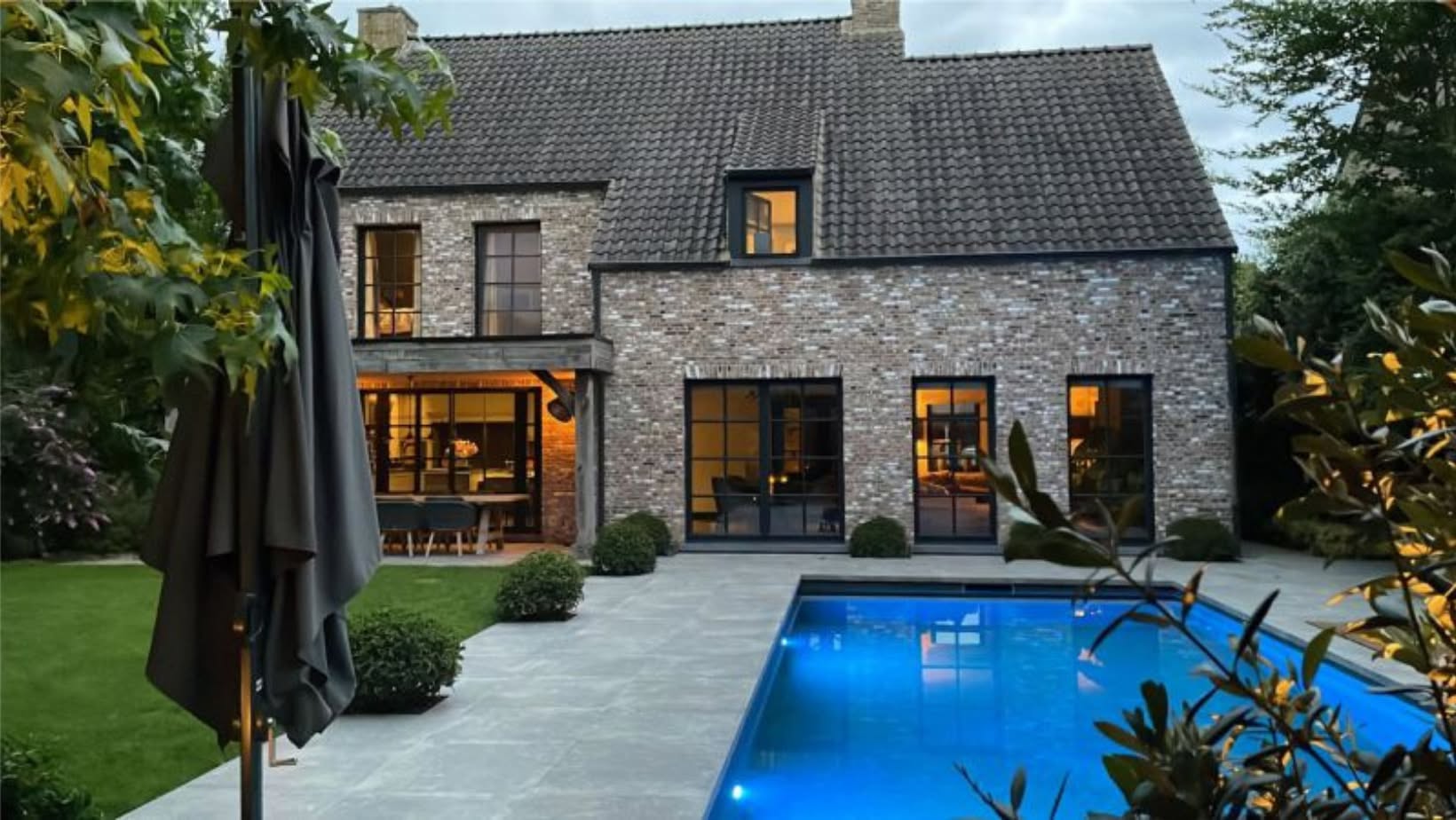 Moderne bakstenen woning met grote ramen, verlichte patio en blauwe zwembad in een groene tuin.