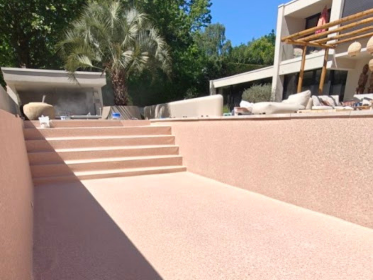 Zonovergoten zwembad met beige muur, ligstoelen en palmbomen tegen blauwe lucht.